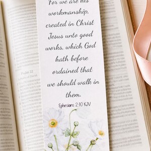 Könnte beinhalten: Ein weißer Lesezeichen mit floralem Design und dem Bibelvers Epheser 2:10 KJV. Das Lesezeichen liegt in einem offenen Buch, mit einem goldenen Stift und einem rosa Band.