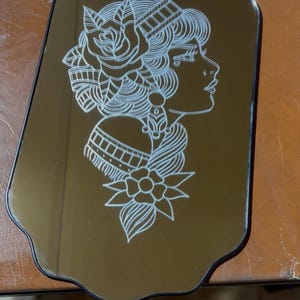 Może przedstawiać: Dekoracyjne lustro o unikalnym kształcie z szczegółowym białym rysunkiem linii profilu kobiety, ozdobione elementami kwiatowymi i ozdobnymi. Lustro wisi na łańcuszku.