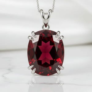 May include: A silver pendant necklace featuring a large, oval-cut, deep red gemstone. The gemstone is set in a silver claw setting and hangs from a delicate silver chain. The pendant is displayed against a white background.