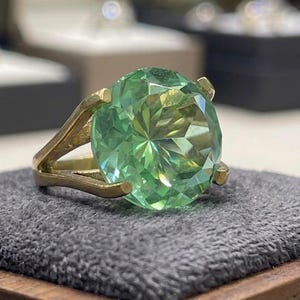 May include: A gold ring featuring a large, faceted, light green gemstone. The ring's band has a split design, and the gemstone is set with four prongs. The ring is displayed on a gray surface.