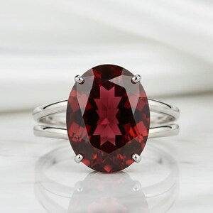 May include: A silver ring with a large, oval-cut, deep red gemstone. The gemstone is the focal point, held by four prongs. The ring has a double-band design, adding a modern touch to the classic gemstone ring.