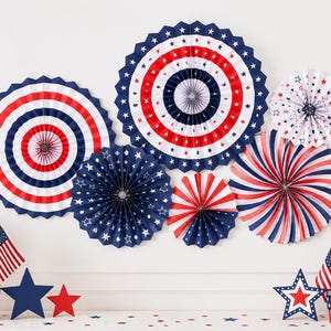 Pode incluir: Uma exibição patriótica com leques de papel vermelhos, brancos e azuis com desenhos de estrelas e listras. O arranjo inclui decorações em forma de estrela, bandeiras americanas em miniatura e arranjos florais.
