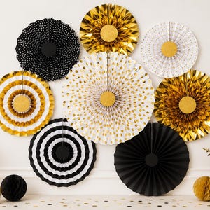 Puede incluir: Un conjunto de abanicos decorativos de papel en dorado, negro y blanco, dispuestos sobre una pared blanca. Los abanicos presentan diversos diseños, incluyendo lunares y rayas. Los detalles dorados y los elementos decorativos completan la exhibición festiva.