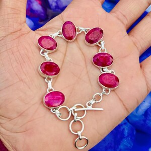Puede incluir: Una pulsera de plata con piedras preciosas rojas ovaladas. Las piedras están engarzadas en eslabones de plata individuales, formando la pulsera. Tiene un cierre de palanca. La pulsera se muestra en una mano.