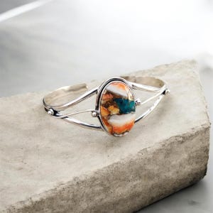 Könnte beinhalten: Ein silbernes Armreif-Armband mit einem ovalen, mehrfarbigen Stein. Der Stein hat orange, weiße und türkisfarbene Farbtöne. Das Armband hat ein schlichtes Design mit geteiltem Band und kleinen dekorativen Akzenten.