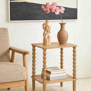 May include: A light wood side table with a lower shelf and turned legs. A vase with a pink orchid and a wooden sculpture sit on top. A stack of books rests on the lower shelf. A beige armchair is to the left.