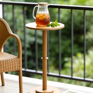 May include: A wooden side table with a round top supports a glass pitcher filled with iced tea and mint. The table has a fluted column and a circular base. A wicker chair and a stack of books are nearby.