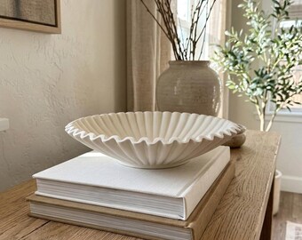 May include: A shallow, off-white ceramic bowl with a scalloped, fluted edge. The bowl is resting on top of two stacked hardcover books, one with a white cover and one with a tan cover, positioned on a light-colored wooden surface.