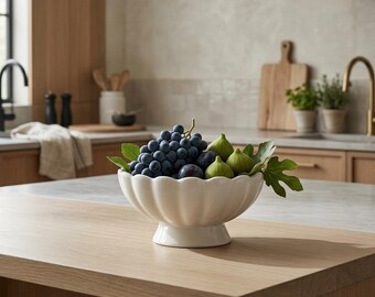 May include: A white ceramic pedestal bowl with a scalloped, fluted rim design. The bowl is filled with a cluster of dark purple grapes and several small, light green figs, accented by a few green fig leaves tucked among the fresh fruit.