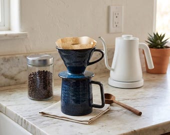 May include: A dark blue ceramic coffee dripper sits atop a matching blue mug, with a light brown paper filter inside the dripper. A clear glass jar filled with coffee beans, a white electric gooseneck kettle, and a wooden spoon are nearby.