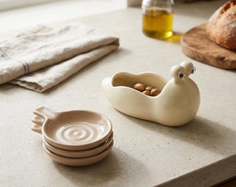 May include: A cream-colored ceramic snail-shaped bowl with googly eyes and a stack of three small, round, light brown ceramic dishes with a concentric circular pattern on the interior base, designed for holding small food items or kitchen trinkets.
