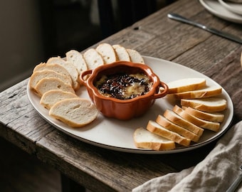 May include: A round, orange, pumpkin-shaped ceramic dish filled with a dark, melted cheese and bacon dip, centered on a large white oval platter surrounded by numerous slices of toasted white baguette bread arranged in two semi-circles.