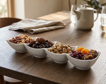 May include: A white ceramic four-section serving dish arranged in a linear row, filled with assorted snacks including almonds, cashews, chocolate-covered treats, pistachios, dried cranberries, dried apricots, and stuffed dates.