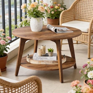 May include: A round, wooden coffee table with a lower shelf, holding books and a tray. A white vase with peach-colored flowers sits on top. Wicker chairs and potted plants surround the table, creating a cozy outdoor setting.