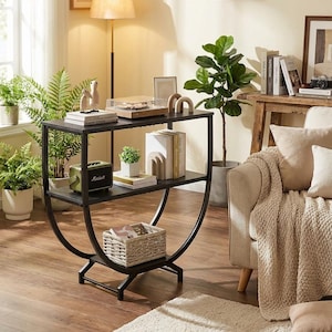 May include: A black metal console table with two shelves, featuring a curved design. The shelves hold a record player, books, a small plant, and a woven basket. The room is decorated with plants, a floor lamp, and a beige sofa.