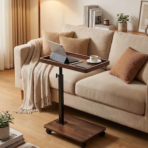 May include: A brown, adjustable laptop table with a black base and wheels. A laptop, a cup of coffee, and a tablet are on the table. The table is next to a beige sofa with decorative pillows.
