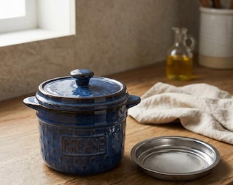 May include: A deep blue ceramic grease container with a matching lid and two side handles. The front of the container features the embossed text "GREASE" surrounded by a subtle dot pattern. A separate circular stainless steel mesh strainer sits nearby.