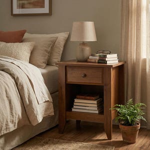 May include: A wooden nightstand with a drawer and lower shelf, holding books. A beige lamp with a fabric shade sits on top, next to a stack of books and eyeglasses. A small potted fern is on the floor. The room has a neutral color palette.