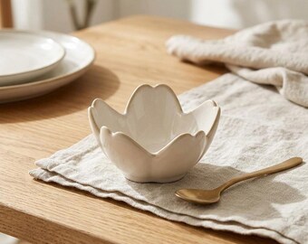 May include: A small, off-white ceramic bowl shaped like a five-petaled flower, accompanied by a slender, metallic gold-toned spoon. The bowl features smooth, curved edges and a glossy finish, resting on a textured, light-colored fabric surface.