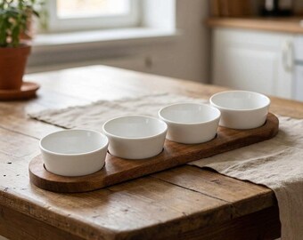 May include: A set of four small, round, white ceramic bowls arranged in a row on a long, narrow, dark brown wooden serving tray. The bowls have smooth, plain surfaces and are designed to sit securely within circular indentations in the wood.