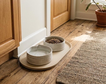 May include: Two round, off-white ceramic bowls with a ribbed, textured exterior sit on an oval, matching off-white tray. One bowl contains dry brown pet food kibble, while the other bowl is filled with clear water, creating a set for pets.