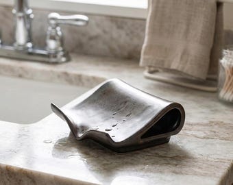 May include: A dark brown, curved ceramic soap dish with a smooth, glossy finish. The dish features a unique, folded-over design that creates a hollow space underneath, with several small water droplets visible on its concave surface.