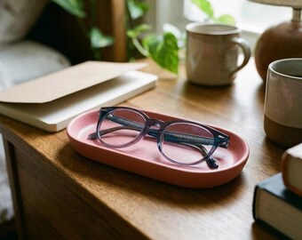 May include: A pair of blue-tinted eyeglasses with dark frames resting inside a smooth, oval-shaped, matte pink ceramic tray. The tray has raised edges and a solid, uniform color, providing a simple surface for the glasses.