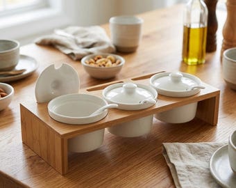 May include: A set of three white ceramic bowls with matching lids and small spoons, arranged in a rectangular bamboo stand. The bowls are designed for serving condiments or spices, featuring a clean, smooth finish and a simple, functional structure.