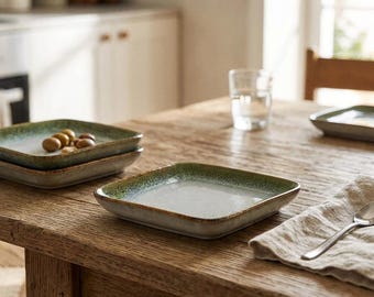 May include: A set of square ceramic plates with a speckled off-white center and a gradient green rim. The plates have a low profile and slightly raised edges. One plate is empty, while another holds a small cluster of brown olives.