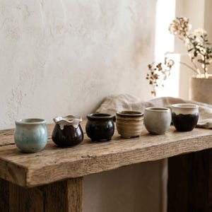 May include: A collection of six small ceramic cups arranged in a row. The vessels feature varied finishes including light blue, dark brown with a white drip glaze, solid black, tan with a dark rim, matte grey, and a two-tone brown and white design.