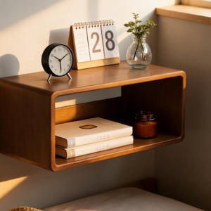 May include: A floating wooden shelf with a minimalist design. It holds a black alarm clock, a calendar displaying the date 28, a small glass vase with greenery, two books, and a candle in a jar. The shelf is mounted on a wall.