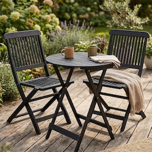 May include: A black outdoor bistro set with two folding chairs and a round table. Two mugs and a folded cloth sit on the table. A tan blanket is draped over one chair. The set is on a wooden deck, with greenery in the background.