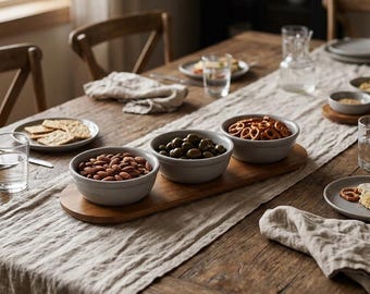 May include: A wooden serving tray holds three round, light gray ceramic bowls filled with almonds, green olives, and pretzels. The tray sits on a long, textured beige linen runner atop a rustic wooden dining table with plates and glasses.