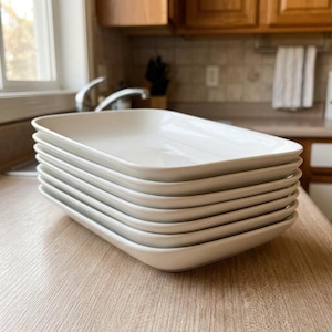May include: A stack of six rectangular white ceramic serving platters with rounded corners and slightly raised edges. The dishes are smooth, glossy, and uniform in size, arranged neatly in a vertical pile on a flat surface.