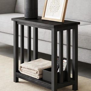 May include: A black wooden side table with a framed picture and a smart speaker on top. The table has a lower shelf with a folded blanket and a small black box with remote controls. The table is on a light-colored rug.