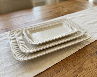 May include: Two rectangular off-white ceramic serving platters stacked on top of each other. Each platter features a raised, textured border with a repeating pattern of vertical lines and small dots along the outer edge of the rim.