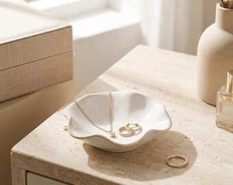 May include: A white ceramic trinket dish with a wavy, scalloped edge. Inside the dish rests a delicate gold chain necklace with a small pendant and two small gold hoop earrings. A single gold band ring sits on the surface nearby.