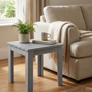 May include: A small, square, gray wooden side table with a slatted top. A potted fern, a mug, and a book rest on the table. The table is next to a beige sofa and a window with beige curtains. The table is approximately 20 inches wide.