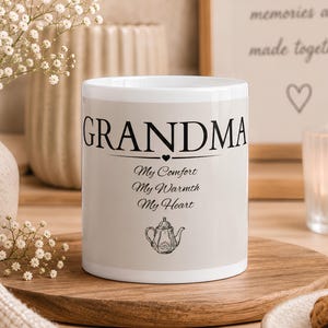 Op de afbeelding: Een witte keramische mok met de zwarte tekst GRANDMA, gevolgd door de woorden My Comfort, My Warmth, My Heart en een kleine illustratie van een theepot. De mok heeft een gladde afwerking en staat op een rond houten oppervlak.