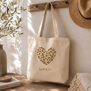 Könnte beinhalten: Eine cremefarbene Stofftasche aus Segeltuch mit einem braunen Herzmotiv im Leopardenmuster auf der Vorderseite. Unter dem Herz ist der Text "Inspiration by Love" in einer dunklen, dünnen Schreibschrift aufgedruckt. Die Tasche hat zwei Tragegriffe.