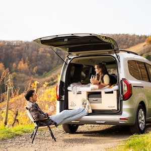 May include: A silver van with its rear door open, showing a bed and storage. A person sits in a folding chair outside, while another is inside. The scene is set in a vineyard with autumn foliage, with a backdrop of hills.