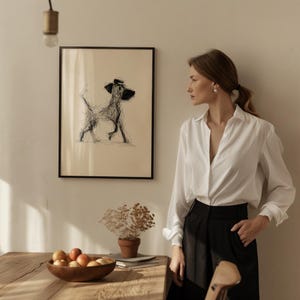 May include: A framed black and white sketch of a dog with a hat. The artwork hangs on a beige wall. A wooden table holds a wooden bowl of fruit, a small potted plant, and a stack of books. A woman in a white shirt and black pants stands nearby.