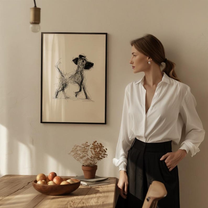 May include: A framed black and white sketch of a dog with a hat. The artwork hangs on a beige wall. A wooden table holds a wooden bowl of fruit, a small potted plant, and a stack of books. A woman in a white shirt and black pants stands nearby.