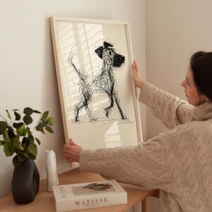 May include: A framed art print featuring a black and white sketch of a dog. The artwork is displayed in a light-colored wooden frame. The dog is depicted in a loose, scribbled style, with visible lines and a playful pose. The background is a neutral beige.