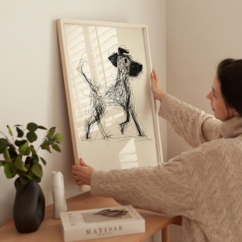 May include: A framed art print featuring a black and white sketch of a dog. The artwork is displayed in a light-colored wooden frame. The dog is depicted in a loose, scribbled style, with visible lines and a playful pose. The background is a neutral beige.