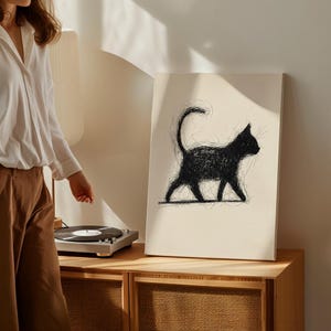 Pode incluir: Impressão artística de gato preto em tela. A obra apresenta um design minimalista e esboçado de um gato em perfil, andando com a cauda curvada. O gato é renderizado em preto sobre um fundo creme. A tela é exibida em um armário de madeira.
