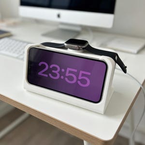 Könnte beinhalten: Eine weiße Ladestation mit einer schwarzen Apple Watch und einem Smartphone, das die Uhrzeit 23:55 in Lila anzeigt. Die Station steht auf einem weißen Schreibtisch, im Hintergrund ein Computermonitor.