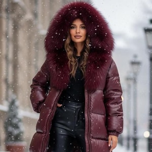 Puede incluir: Un abrigo acolchado burdeos con capucha ribeteada de piel. El abrigo llega hasta la rodilla y tiene un acabado brillante. La modelo lleva pantalones negros y un top negro. Está nevando en el fondo.