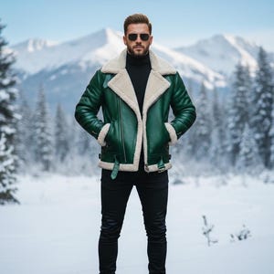 May include: A green leather jacket with a cream-colored shearling lining and collar. The jacket has a classic aviator style with a zip closure, pockets, and buckle details. The person in the image is wearing black pants and sunglasses, standing in a snowy landscape.