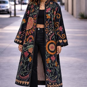 Puede incluir: Un abrigo largo de terciopelo azul oscuro con bordados florales coloridos. El abrigo presenta un cuello mandarín y un diseño largo y fluido. El bordado incluye flores y hojas naranjas, rosas y verdes. El abrigo se usa sobre pantalones negros y una parte superior negra.